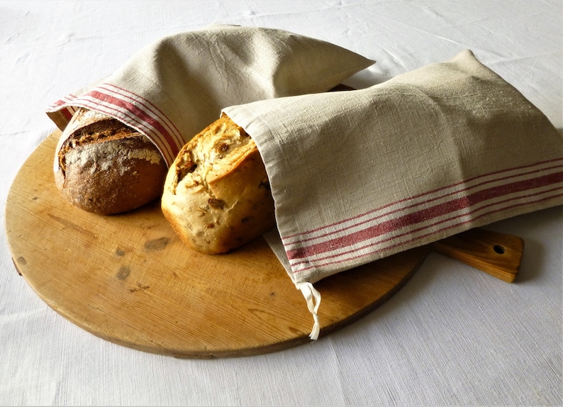 Op de afbeelding: Twee broden op een houten snijplank, bedekt met linnen doeken. De doeken zijn beige met rode strepen. E&eacute;n brood is donkerbruin, het andere is goudbruin.