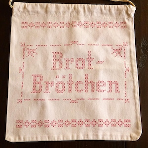 Op de afbeelding: Cr&egrave;mekleurige zak met trekkoord en rode kruissteekborduurwerk. De tas heeft de Duitse woorden "Brot-Br&ouml;tchen" in een decoratief lettertype, omgeven door gestikte randen en bloemaccenten. Een praktisch en charmant item.