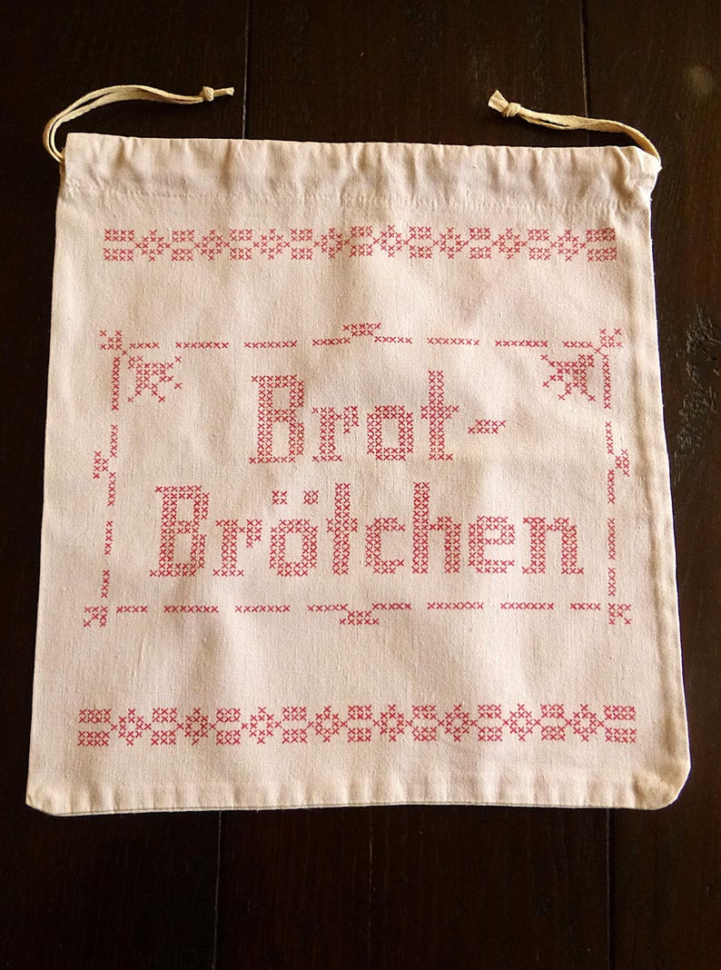 Op de afbeelding: Cr&egrave;mekleurige zak met trekkoord en rode kruissteekborduurwerk. De tas heeft de Duitse woorden "Brot-Br&ouml;tchen" in een decoratief lettertype, omgeven door gestikte randen en bloemaccenten. Een praktisch en charmant item.