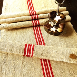 May include: Beige linen table runner with three vertical red stripes. Rolled linen napkins are stacked on top of the runner. A two-tiered silver serving tray holds biscuits and chocolates. A small wooden tag is attached to the runner.