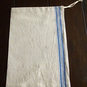 Op de afbeelding: Een rechthoekige beige linnen trekkoordtas met een verticaal blauw gestreept ontwerp aan de rechterkant. De tas heeft een natuurlijke, licht gekreukelde textuur en een cr&egrave;mekleurige trekkoordsluiting.