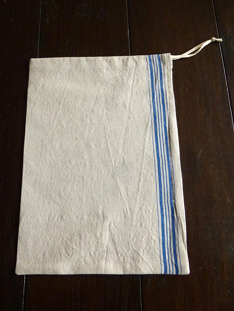 Op de afbeelding: Een rechthoekige beige linnen trekkoordtas met een verticaal blauw gestreept ontwerp aan de rechterkant. De tas heeft een natuurlijke, licht gekreukelde textuur en een cr&egrave;mekleurige trekkoordsluiting.