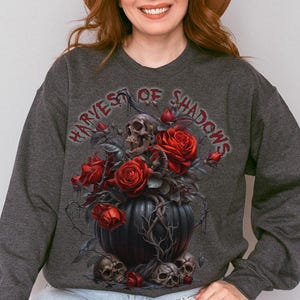 May include: Dark gray sweatshirt featuring a graphic of skulls, red roses, and the text "HARVEST OF SHADOWS." The design is centered on the front of the long-sleeved top. The person in the image is wearing a brown hat.