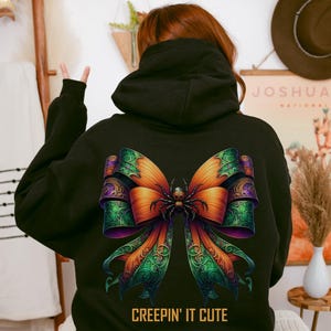 May include: Black hooded sweatshirt featuring a colorful bow design with a spider in the center. The bow has orange, green, and purple accents. The text "CREEPIN' IT CUTE" is printed in gold at the bottom of the sweatshirt. The sweatshirt is a Halloween-themed apparel item.