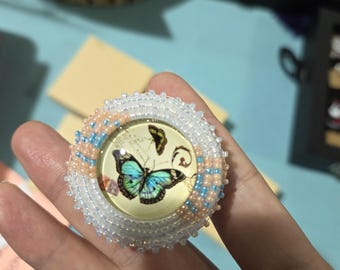 May include: A round, beaded brooch featuring a butterfly design. The brooch has a central image of a butterfly with blue and green wings, surrounded by a border of white, peach, and blue beads. The brooch is held in a hand.