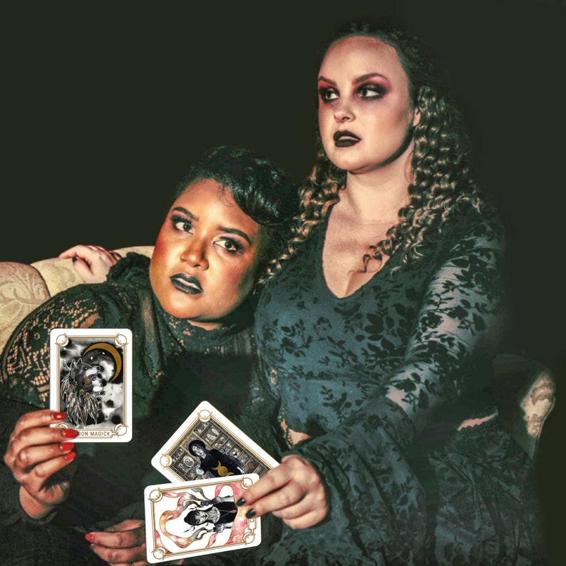 A black and white witch wear dark vamp gothic lace Victorian dresses and witchy makeup and are holding oracle cards, like the tarot as they sit on a vintage beige tan couch