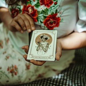 May include: A tarot card with the word "ABUNDANCE" at the bottom. The card features an illustration of a woman holding a fruit, with a gold circle behind her. The card is held in front of a floral patterned background.