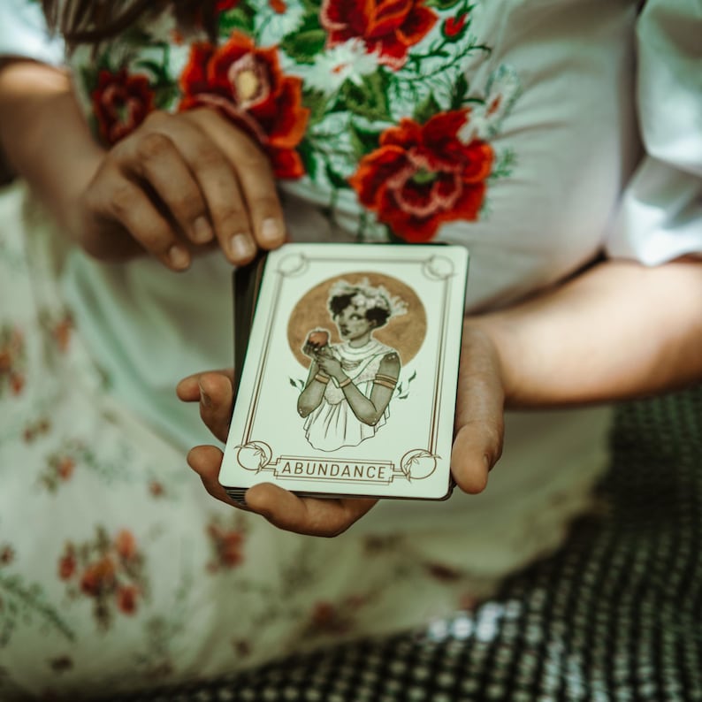 May include: A tarot card with the word "ABUNDANCE" at the bottom. The card features an illustration of a woman holding a fruit, with a gold circle behind her. The card is held in front of a floral patterned background.