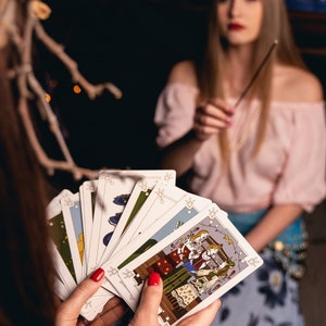 A woman wearing a head crown of branches holds a set of divination cards while her friend waves incense. The card shown in her hand shows Koledari, Slavic women wearing their traditional Russian, Polish, Ukrainian dress singing during Christmas time
