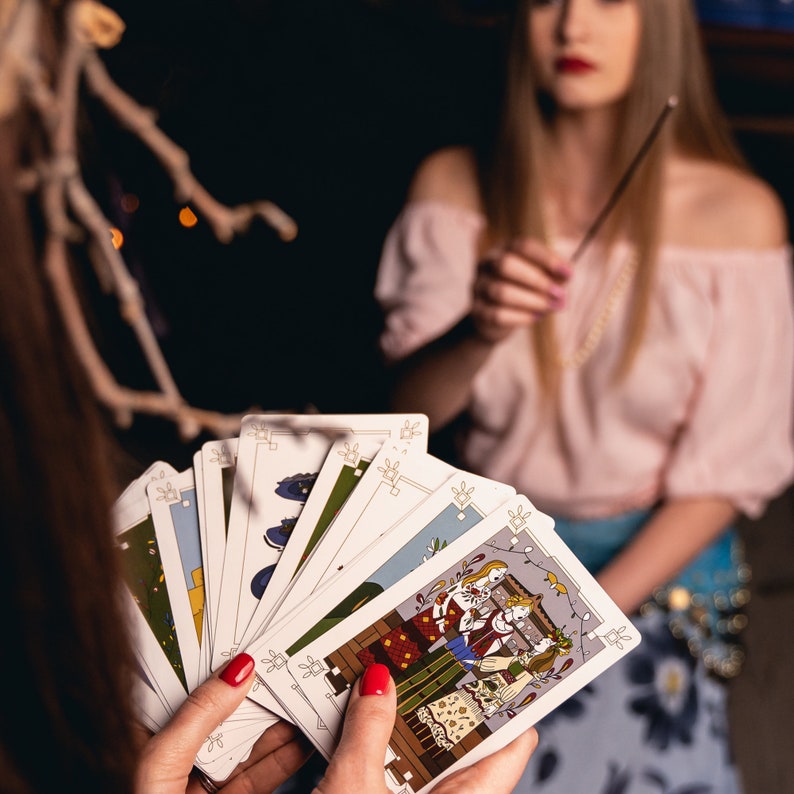 A woman wearing a head crown of branches holds a set of divination cards while her friend waves incense. The card shown in her hand shows Koledari, Slavic women wearing their traditional Russian, Polish, Ukrainian dress singing during Christmas time