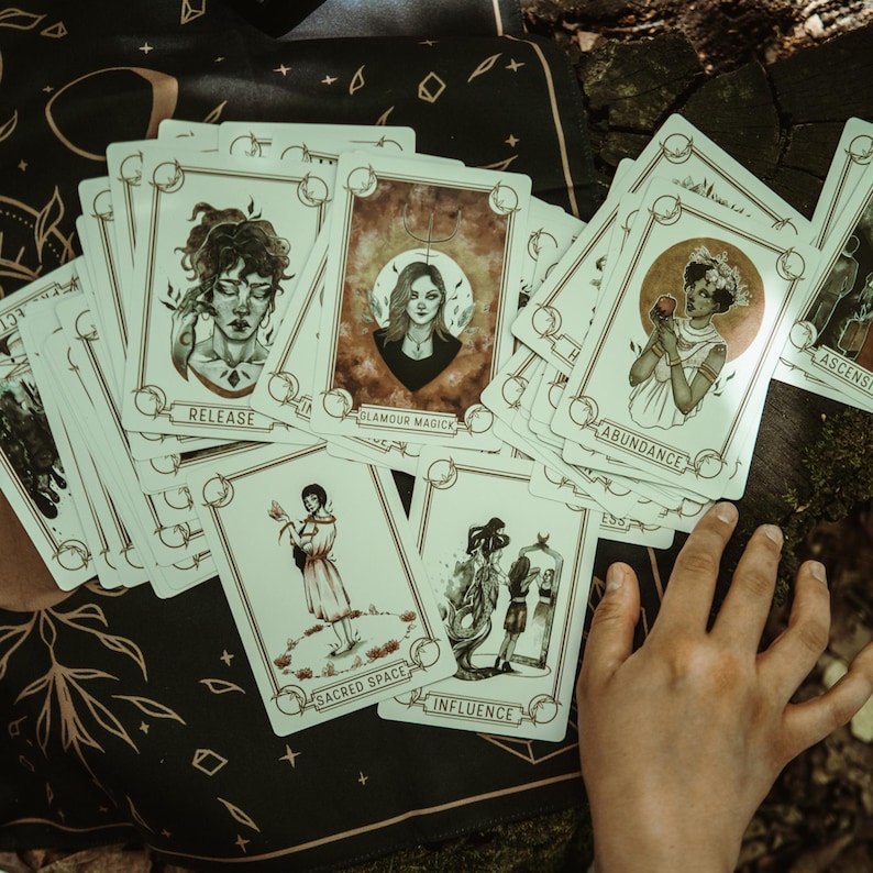 May include: A collection of tarot cards featuring illustrations of women and symbolic imagery. Each card has a unique design with titles like "Release," "Glamour Magick," and "Abundance." The cards are arranged on a dark cloth with a celestial pattern.