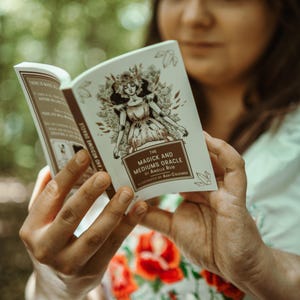 May include: A book titled "The Magick and Mediums Oracle" is held open, revealing illustrations and text. The cover features a sepia-toned drawing of a woman. The book's pages display additional text and illustrations, suggesting a guide or reference.