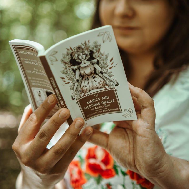 May include: A book titled "The Magick and Mediums Oracle" is held open, revealing illustrations and text. The cover features a sepia-toned drawing of a woman. The book's pages display additional text and illustrations, suggesting a guide or reference.
