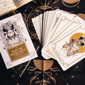 An oracle deck guidebook with a woman wearing a crystal crown laying on a bed of crystals with hands on her lies on a witchy cloth next to Magick and Mediums oracle cards. The abundance card with a young black woman holding an apple sits on top.