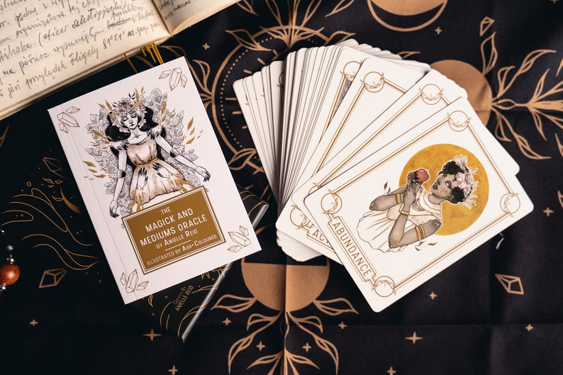 An oracle deck guidebook with a woman wearing a crystal crown laying on a bed of crystals with hands on her lies on a witchy cloth next to Magick and Mediums oracle cards. The abundance card with a young black woman holding an apple sits on top.