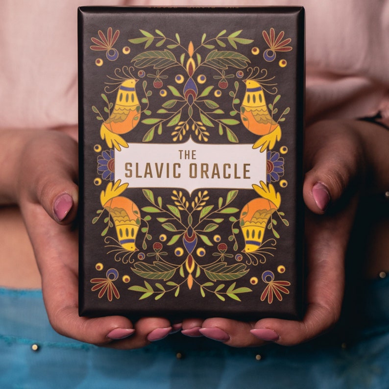 A Polish woman wearing a pink top and a blue skirt is holding The Slavic Oracle deck box which is decorated with Slavic imagery like birds and folk-art flowers of bright yellow, green, blues and reds over a dark background.
