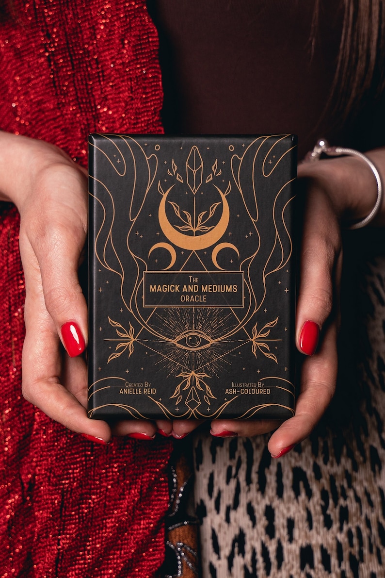 May include: A black oracle card deck titled "The Magick and Mediums Oracle" with gold illustrations of moons, crystals, and an eye. The deck is held in hands with red nail polish, against a red sequined top and leopard print background.