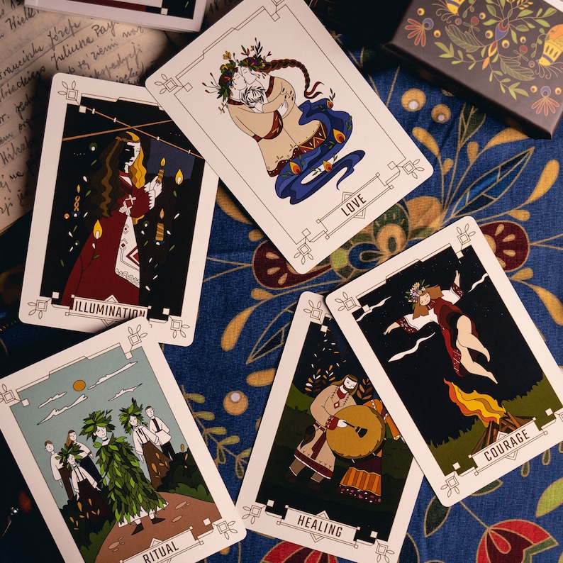 A few cards from The Slavic Oracle are on a blue cloth decorated with traditional Slavic folk floral art. The cards show the Kust or Dodola ritual, a Slavic priest or Volkh playing a healing drum and a girl and couple on Kupala, the Summer Solstice
