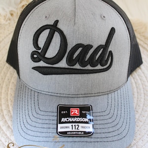 Può includere: Cappellino da camionista grigio con la scritta "Dad" ricamata in nero. Il cappellino ha un retro in rete nera e una visiera curva. Una toppa Richardson 112 Trucker è visibile sul davanti.