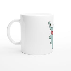 Könnte beinhalten: Ein weißer Keramikbecher mit einer Illustration eines hellblauen, amöbenartigen Wesens. Das Wesen hat ein gelbes Auge mit schwarzer Pupille und auffällige rote Lippen. Das Motiv befindet sich auf der Seite des glatten, zylindrischen Bechers.