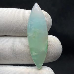 Könnte beinhalten: Ein polierter, länglicher Edelstein mit spitzer Spitze. Der Stein zeigt einen Farbverlauf, der von einem durchscheinenden Weiß oben zu einem sanften Grün an der Basis übergeht. Wispige, wolkenartige Einschlüsse sind im Stein sichtbar.
