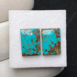 Op de afbeelding: Twee rechthoekige turkoois edelstenen met bruine matrixpatronen. De stenen liggen op een wit oppervlak en tonen hun levendige blauwgroene kleur en unieke adering.