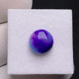 May include: A round, polished gemstone with a swirling pattern of deep purple and light blue hues. The stone is set on a white, square surface, creating a striking contrast. The gemstone's smooth surface reflects light, highlighting its vibrant colors.