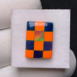 Op de afbeelding: Een rechthoekig object met een dambordpatroon van afwisselend oranje en marineblauwe vierkanten. Het middelste vierkant heeft een iriserend, glinsterend effect. Het object staat tegen een witte achtergrond.