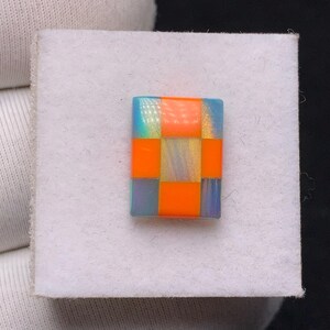 Op de afbeelding: Een klein, rechthoekig object in mozaïekstijl met een dambordpatroon. De vierkanten wisselen af tussen fel oranje en iriserende blauwgroene tinten. Het object staat tegen een witte, getextureerde achtergrond.