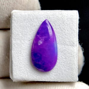 May include: A teardrop-shaped gemstone with a polished surface. The stone displays a gradient of purple hues, transitioning from deep violet to lighter shades. The gemstone is set against a white background.