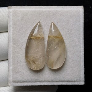May include: Two teardrop-shaped gemstones with a translucent, light brown hue. Fine, golden, needle-like inclusions are visible within the stones. The gems are set against a white, textured background, with a black backdrop.