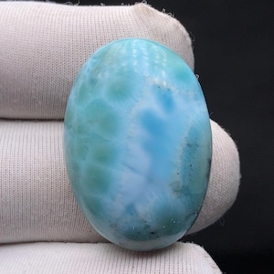 May include: Oval-shaped larimar gemstone, showcasing a blend of light and deep blues with white marbling. The polished surface reflects light, highlighting the stone's natural patterns. The gemstone is held against a neutral background.