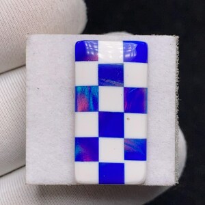 Op de afbeelding: Een rechthoekig object met een dambordpatroon van witte en blauwe vierkanten. De blauwe vierkanten hebben iriserende kleurwervelingen. Het object ligt op een wit oppervlak en heeft afgeronde randen.