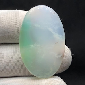 Op de afbeelding: Een ovaalvormige edelsteen met een gepolijst oppervlak. De steen heeft een mix van kleuren, waaronder groen, blauw en wit, wat een wolkachtige, etherische uitstraling geeft. De edelsteen wordt tegen een witte stoffen achtergrond gehouden.