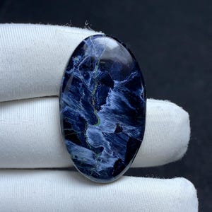 May include: An oval-shaped pietersite gemstone cabochon featuring a polished surface with deep navy blue and black tones, accented by swirling, chatoyant streaks of light blue and thin, vibrant green mineral inclusions throughout the stone.