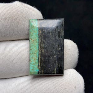 Puede incluir: Una gema rectangular con una superficie pulida. La piedra presenta una vibrante franja turquesa en el lado izquierdo, que contrasta con el patrón oscuro, casi negro, texturizado en el lado derecho. La gema es sostenida por una mano enguantada de blanco.