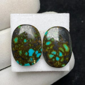 May include: Two oval-shaped turquoise gemstones with a mix of colors. The stones have a dark matrix with patches of bright turquoise and olive green. The stones are set on a white surface, showcasing their natural beauty.