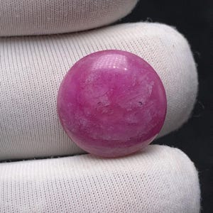Könnte beinhalten: Nahaufnahme eines runden, polierten Edelsteins. Der Edelstein hat eine leuchtende magentafarbene Farbe mit subtilen Farb- und Texturvariationen. Der Stein wird vor einem weißen Stoffhintergrund gehalten.