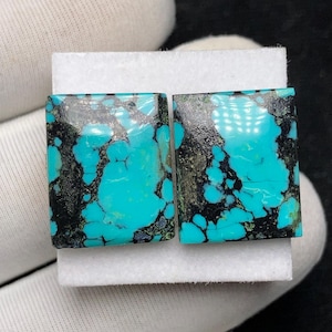 Op de afbeelding: Twee vierkante turkoois edelstenen met een opvallende matrix van zwarte en grijze aderen. De gepolijste stenen tonen een levendige, helder blauwgroene kleur, tegen een witte achtergrond. Deze edelstenen zijn ideaal voor het maken van sieraden.