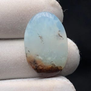 Könnte beinhalten: Ein ovaler Edelstein mit einem wolkigen, himmelblauen und weißen Muster. Der untere Teil weist ein braunes und beiges Erddesign auf, das einen einzigartigen Kontrast erzeugt. Der Stein wird von einem weißen Stoff gehalten.