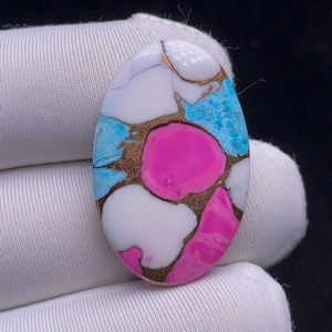May include: An oval-shaped gemstone with a mosaic design. The stone features a combination of white, light blue, and pink sections, separated by a gold-colored matrix. The gemstone is held in a white glove.