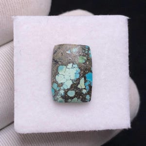 Op de afbeelding: Een rechthoekige turkoois edelsteen met een gevlekt patroon van lichtblauwe, donkerblauwe en grijze tinten. De steen is geplaatst tegen een witte achtergrond, die de natuurlijke kleuren en texturen laat zien. De edelsteen wordt waarschijnlijk gebruikt voor het maken van sieraden.