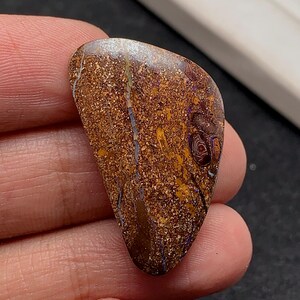 Puede incluir: Ópalo pulido en forma de lágrima con una rica paleta de colores terrosos. La piedra presenta una base marrón moteada con vetas doradas, azules y moradas. La superficie del ópalo tiene una apariencia texturizada única, mostrando patrones naturales.