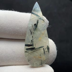 Può includere: Una gemma a forma di goccia con una base trasparente e chiara. La pietra presenta inclusioni a forma di ago verde scuro e alcune macchie giallo dorato. La superficie lucida riflette la luce, evidenziando i motivi interni. La gemma è tenuta contro un tessuto bianco.