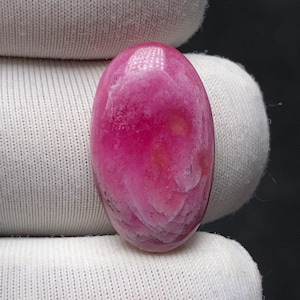 Könnte beinhalten: Ein polierter, oval geformter Edelstein in leuchtendem Rosa. Der Stein zeigt ein marmoriertes Muster mit helleren rosa und weißen Wirbeln, wodurch eine einzigartige visuelle Textur entsteht. Der Edelstein wird vor einem weißen Stoffhintergrund gehalten.