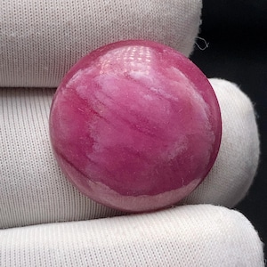 Peut inclure: Une pierre précieuse ronde et polie, d'un rose vif avec des motifs blancs tourbillonnants. La pierre est lisse et réfléchissante, sur un fond de tissu blanc. La pierre mesure environ 2,5 cm de diamètre.