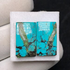 Op de afbeelding: Twee rechthoekige turkoois edelstenen met donkere adering en vlekken groen en grijs. De stenen liggen op een wit oppervlak en tonen hun levendige blauwgroene kleur en natuurlijke patronen. Deze stenen zijn waarschijnlijk voor het maken van sieraden.