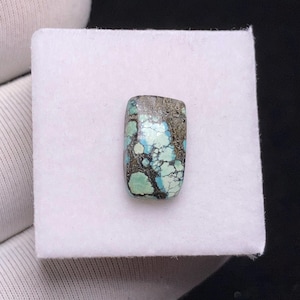 Op de afbeelding: Een rechthoekige turkoois edelsteen met bruine en zwarte aderen. De gepolijste steen ligt op een witte achtergrond. De turkooiskleur is een mix van lichte en donkere tinten.