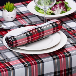 Op de afbeelding: Een wit tafelkleed met een rood, zwart en grijs ruitpatroon. Op het tafelkleed ligt een wit bord met een opgerolde geruite servet. Links van het bord ligt een vork en rechts een mes.