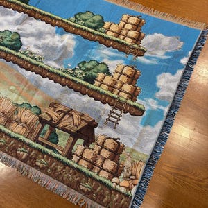 May include: A woven throw blanket featuring a pixelated landscape design with floating grassy islands, stacks of hay bales, and a wooden structure. The scene includes a bright blue sky with white clouds, green bushes, and a brown earth base with fringe.